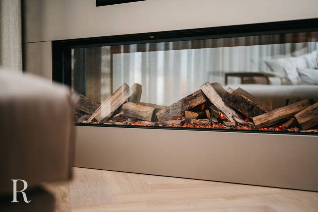 Een moderne binnenhaard met gestapelde houten houtblokken en kunstmatige sintels, omhuld door glas, naast een lichtgekleurde houten vloer en neutraal getint meubilair.