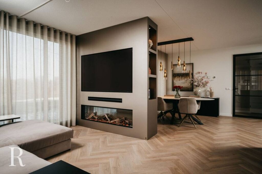 Moderne woon- en eetruimte met een grote tv boven een ingebouwde elektrische open haard op een scheidingsmuur, lichthouten visgraatvloer, zachtbeige meubels en hangende hanglampen boven een eettafel.