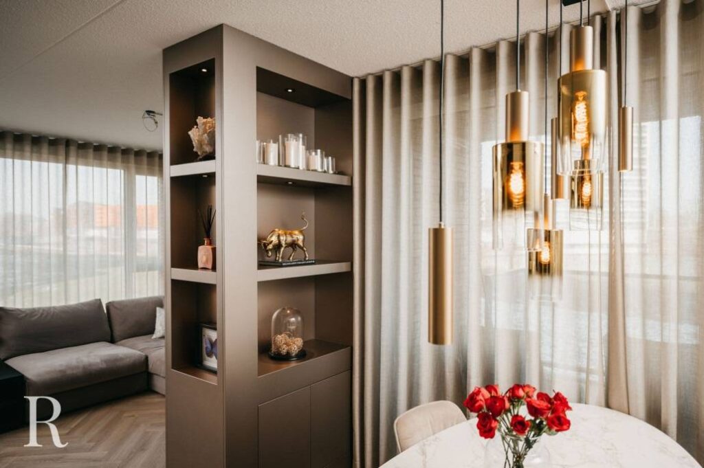 Moderne woon- en eetkamer met beige gordijnen, een grijze sofa, een decoratieve plank met kaarsen en beelden, een ronde eettafel met rode bloemen en gouden hanglampen boven de tafel.