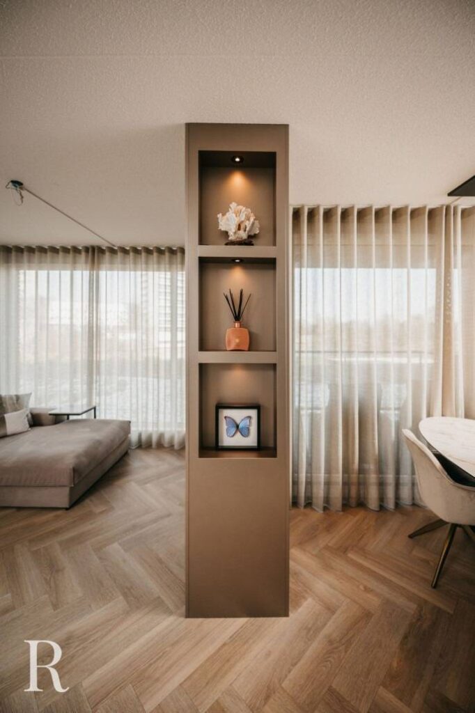Een moderne woonkamer met een ingebouwde schapzuil met een koraalornament, rietverdamper en een ingelijste blauwe vlinder, tegen grote ramen met transparante gordijnen en een houten vloer met visgraatmotief.