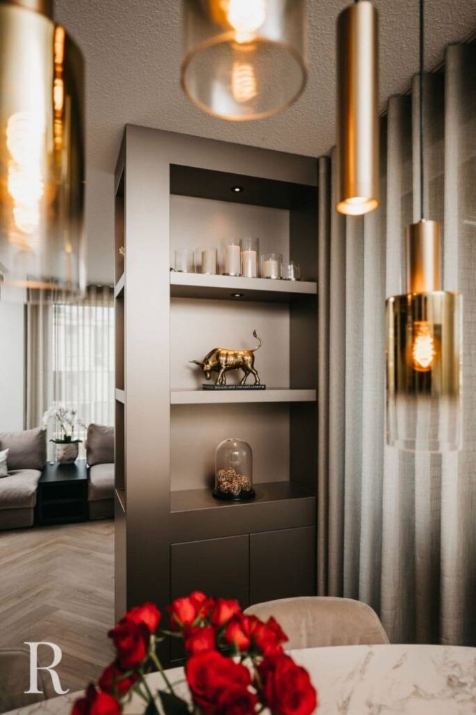 Moderne woonkamer met gouden hanglampen, planken met kaarsen en een gouden stierenbeeldje, transparante gordijnen, marmeren tafel en een boeket rode rozen op de voorgrond. Gezellige zithoek zichtbaar op de achtergrond.