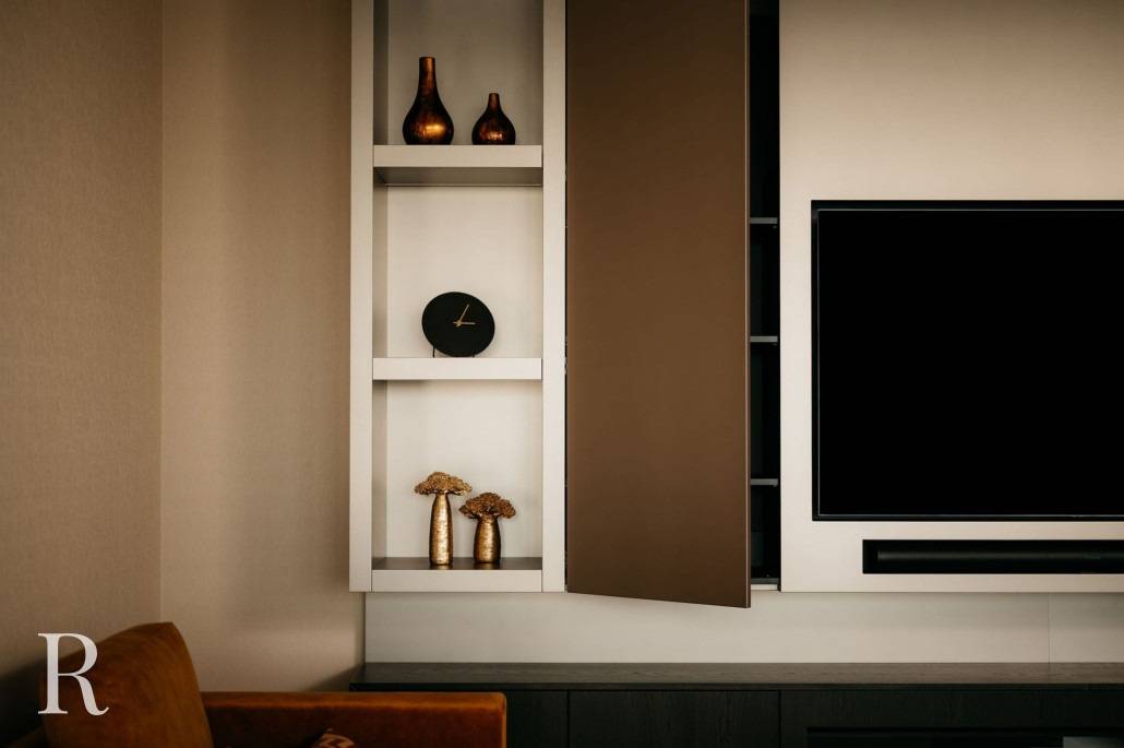 Moderne woonkamer met een strakke Cinewall met een tv aan de muur, een gedeeltelijk open kast, planken met decoratieve vazen, een kleine klok en gedroogde bloemen; overal zachte, warme verlichting en neutrale tinten.