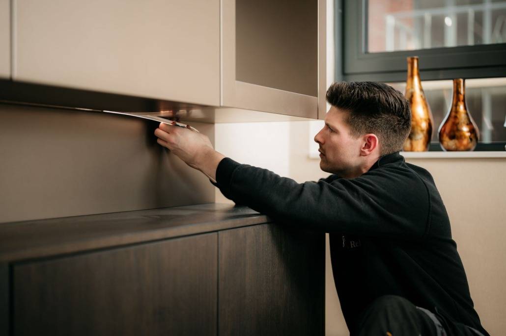 Een man knielt terwijl hij met een meetlint de uitlijning van een wandkast onder een strakke cinemamuur controleert in een moderne, goed verlichte kamer met decoratieve vazen op de vensterbank achter hem.