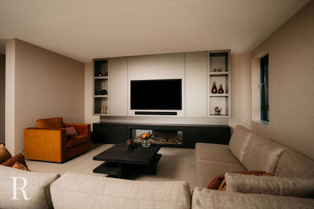 Moderne woonkamer met neutraal gekleurde muren en een grote L-vormige beige bank, een zwarte salontafel, een oranje fauteuil en een ingebouwd cinewall entertainmentcentrum met een flatscreentelevisie en decoratieve planken.