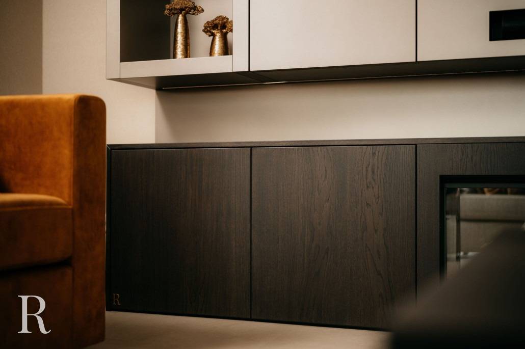 Een moderne woonkamerhoek met een bruin gestoffeerde stoel, donkerhouten kasten en een lichte plank met gouden vazen met gedroogde bloemen. Zachte verlichting accentueert het minimalistische ontwerp, aangevuld met een strakke cinewall voor een elegante touch.