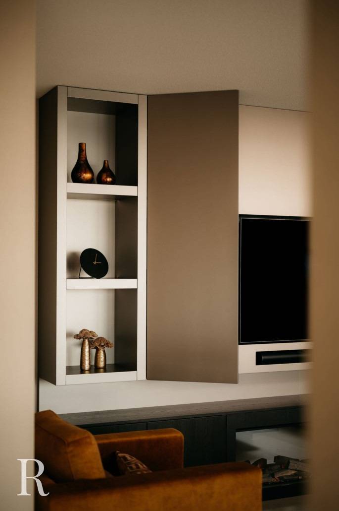 Moderne woonkamerrekken met bronzen vazen, een zwarte klok, droogbloemen, een gemonteerde tv-cinewall en een bruine fauteuil op de voorgrond. De ruimte heeft een minimaal en elegant ontwerp.