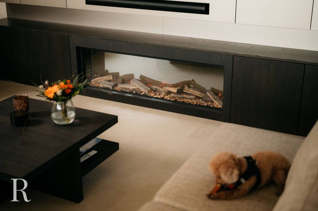 Een klein hondje ligt op een beige sofa in een moderne woonkamer met een cinewall met een elektrische open haard en een donkerhouten salontafel versierd met een vaas bloemen.