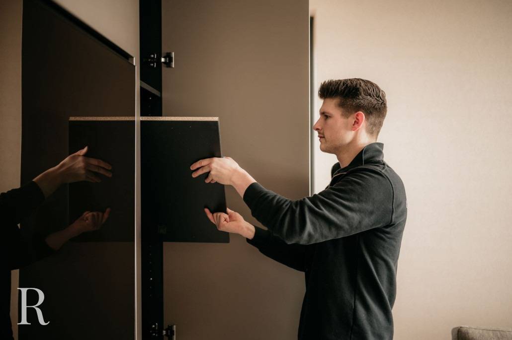 Een man in een zwart overhemd plaatst voorzichtig een plank in een moderne kast, mogelijk om deze voor te bereiden voor een cinewall opstelling. De kastdeur staat open terwijl hij de plank met beide handen bijstelt, tegen neutraal gekleurde muren binnenshuis.