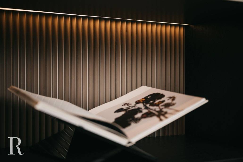 Een open boek met een vage afbeelding rust op een moderne zwarte standaard tegen een gestructureerde, verticaal gelijnde muur die van bovenaf wordt verlicht door warme, zachte verlichting.