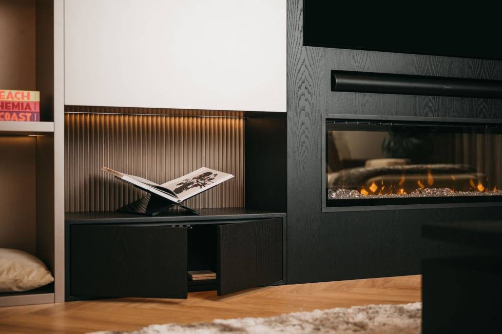 Een moderne woonkamer met een elektrische open haard, een boek op een standaard in een hoekje en boeken op planken. Warme verlichting en houten vloeren creëren een gezellige, eigentijdse sfeer.