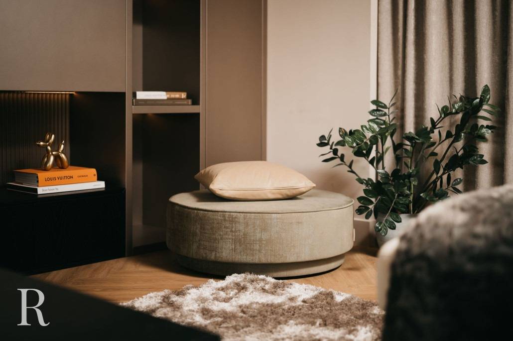 Een gezellige woonkamerhoek met een ronde poef met daarop een beige kussen, een plant in een bladpot, een zacht ruig tapijt, boeken en een gouden beeld van een ballonhond op een kast. Neutrale en warme tinten domineren de ruimte.