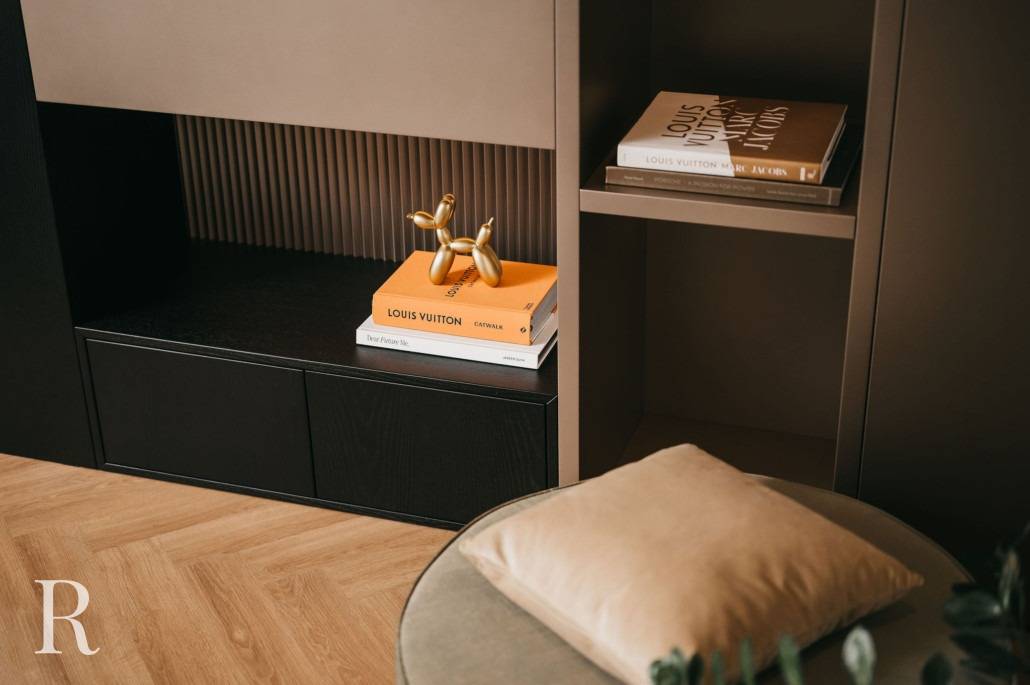 Een stijlvolle binnenplank met gestapelde boeken, waaronder Louis Vuitton, een gouden beeld van een ballonhond en een bijzettafel met een beige kussen op de voorgrond. De vloer is van hout met een visgraatmotief.