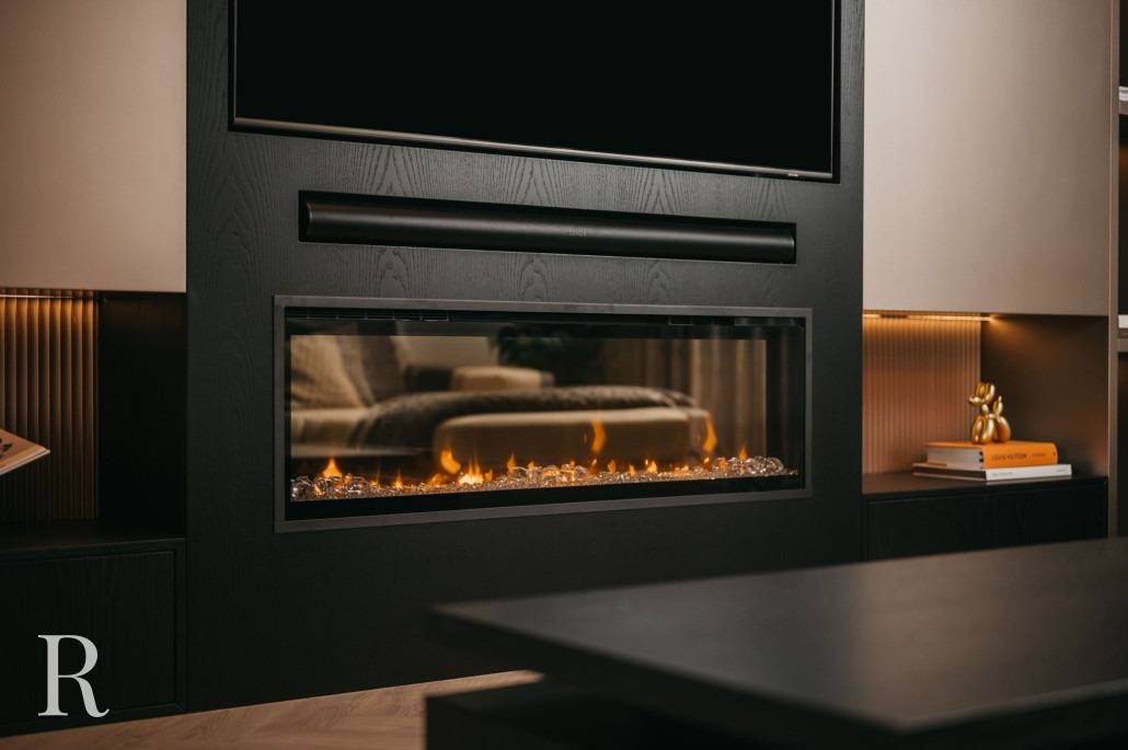 Een moderne woonkamer met een strakke, ingebouwde elektrische open haard met een glazen voorkant onder een gemonteerde tv, omringd door donkere kasten en warme verlichting. Op de voorgrond staat een zwarte salontafel.