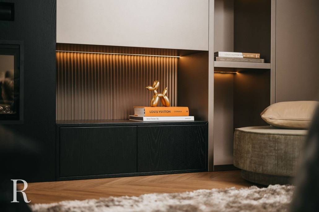 Een moderne woonkamer met een gouden ballonhond sculptuur en Louis Vuitton boeken, met accentverlichting, donkere kasten, een gestructureerd wandpaneel en een ronde poef gedeeltelijk zichtbaar op de voorgrond.