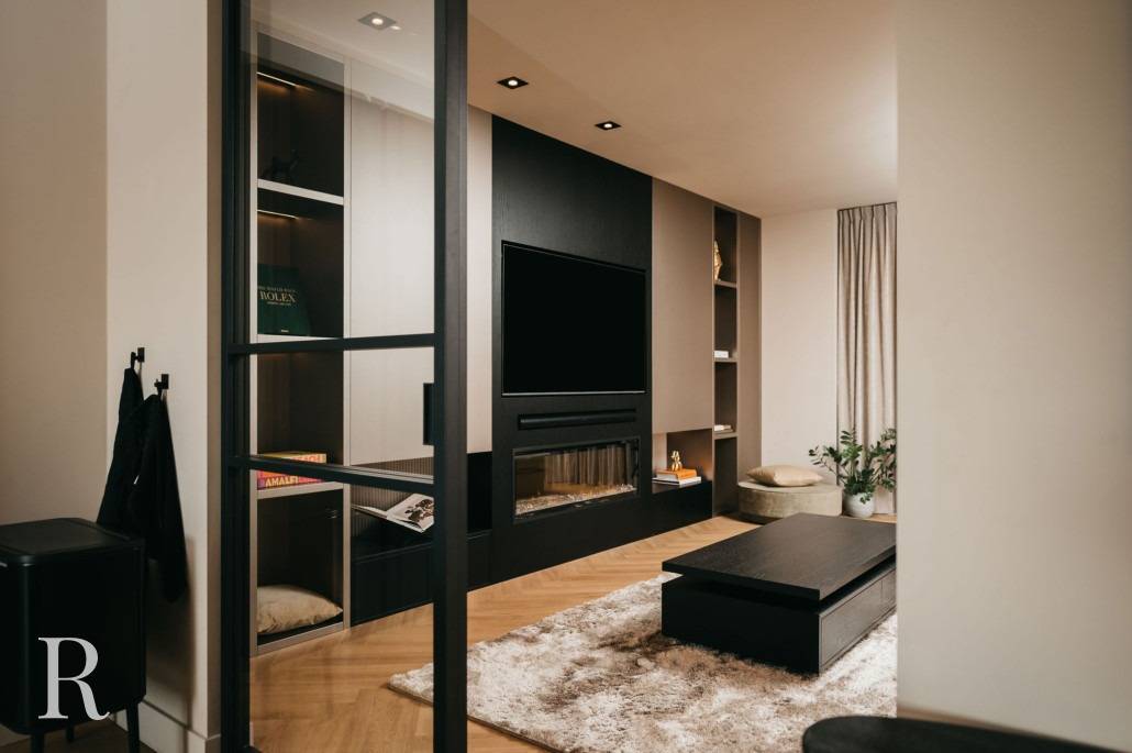 Een moderne woonkamer met een tv aan de muur, ingebouwde planken, een zwarte salontafel, een beige vloerkleed, een plant in de hoek en zachte verlichting. De inrichting is minimaal met neutrale kleuren en houten vloeren.