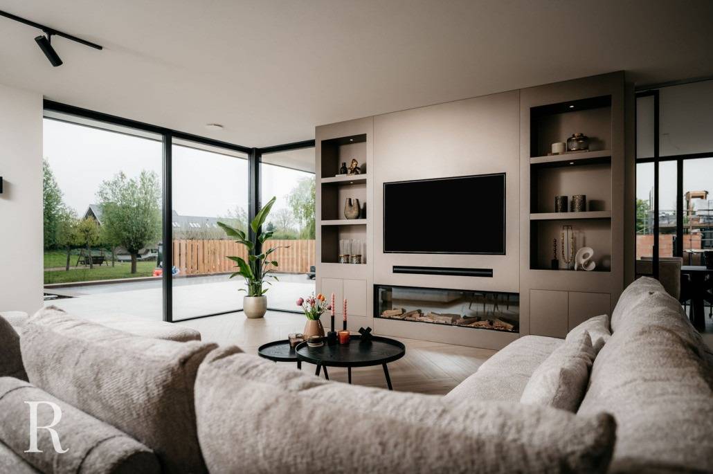 Moderne woonkamer met grote ramen, een beige sofa, ronde salontafel met planten, ingebouwde planken, een flatscreentelevisie en een elektrische open haard. Buitenpatio en groen zichtbaar door de ramen.