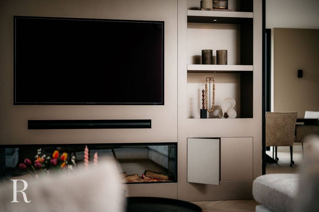 Een moderne muur in de woonkamer is voorzien van een grote flatscreentelevisie, ingebouwde planken met decoratieve voorwerpen, een soundbar en een open haard met glazen wanden. Een vaas met bloemen en een deel van een eethoek zijn ook zichtbaar.