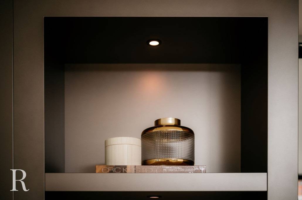 Een plank met een spotlight toont een geribbelde witte pot, een glazen pot met textuur en een gouden deksel en twee gestapelde boeken met zichtbare ruggen in een moderne, minimalistische omgeving. Linksonder staat een grote letter R.