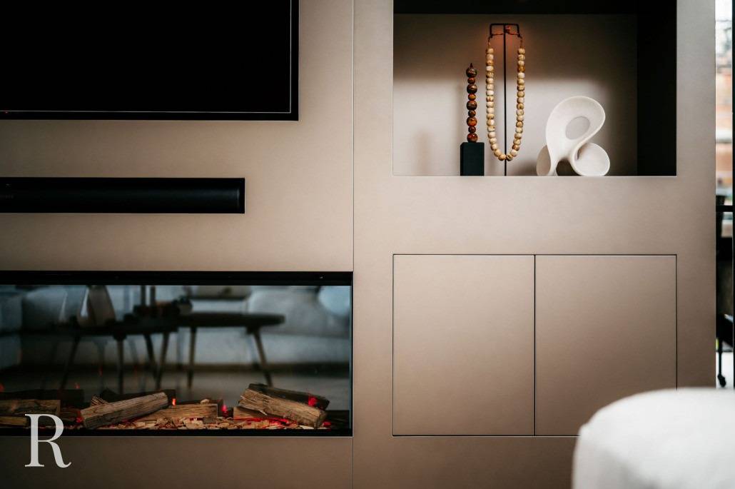 Een moderne woonkamerwand met een ingebouwde tv, open haard met gestapelde houtblokken en een decoratieve nis met een lamp, houten kralen en een wit abstract beeld. Het ontwerp is minimalistisch en eigentijds.