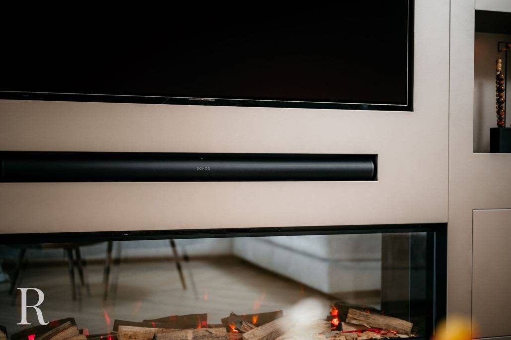 Een moderne woonkamer met een tv aan de muur, een zwarte soundbar eronder en een elektrische open haard met decoratieve houtblokken eronder. De hoek van een stoel en tafel zijn zichtbaar op de achtergrond.