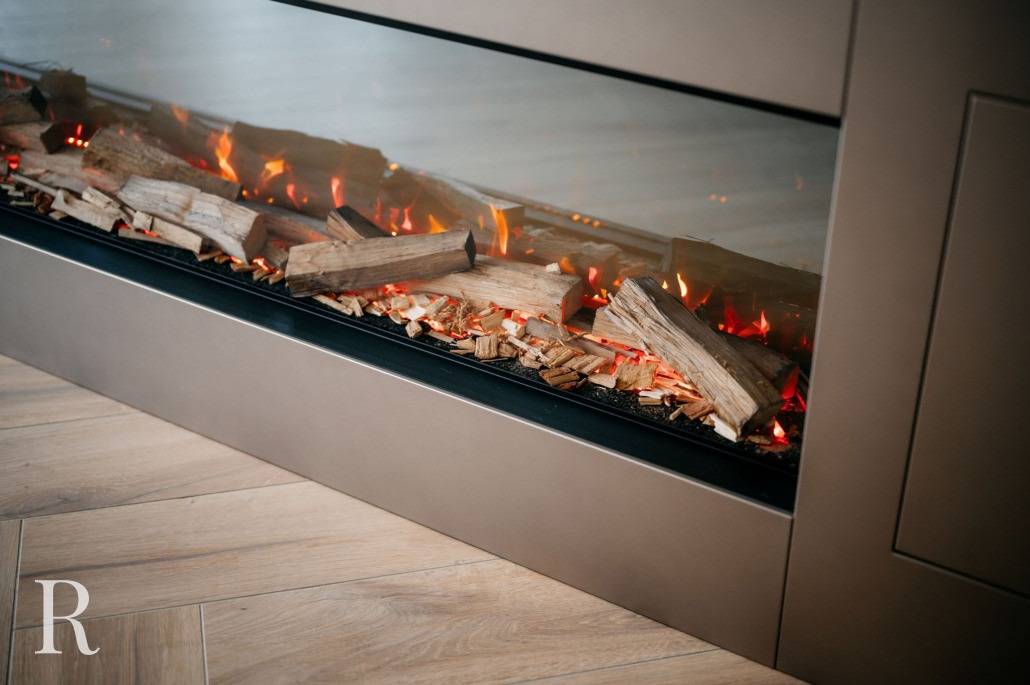 Een close-up van een moderne elektrische open haard met kunstmatige houtblokken en gloeiende oranje vlammen, in een strakke behuizing met glazen frame op een houten vloer.