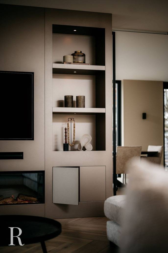 Moderne ingebouwde planken met decoratieve voorwerpen, waaronder vazen, potten en kralen, staan tegen een neutrale beige muur. Links zijn een tv en een deel van een open haard zichtbaar, met een eethoek op de achtergrond.