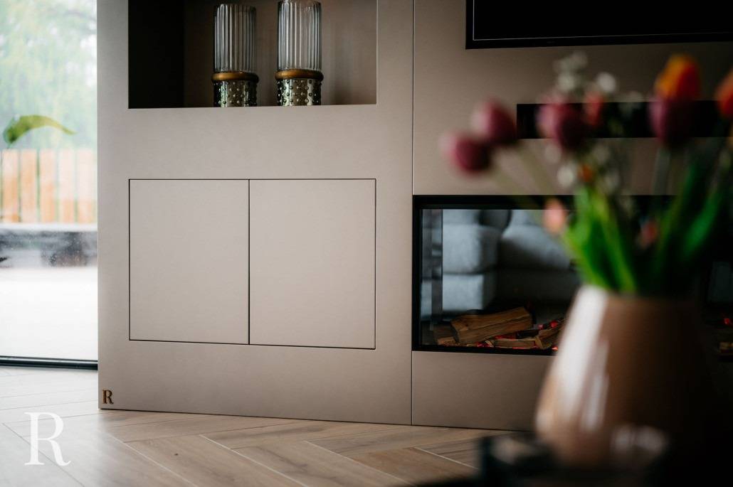 Een moderne woonkamer met een strakke kast, een open haard met gestapeld hout en een vage vaas met kleurrijke tulpen op de voorgrond. Licht stroomt naar binnen door een groot raam, waardoor een neutraal, minimalistisch ontwerp wordt benadrukt.