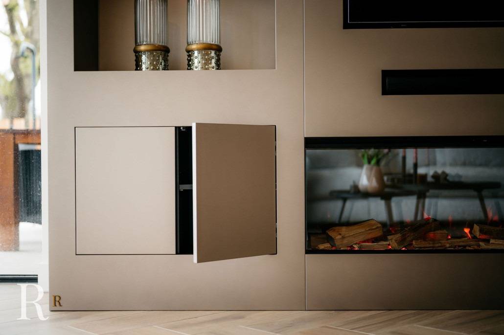 Een modern interieur met een beige wandmeubel met een deurtje dat gedeeltelijk openstaat naast een elektrische open haard met gestapelde houtblokken. Twee decoratieve vazen staan op een bovenste plank. Licht komt binnen door een groot raam.