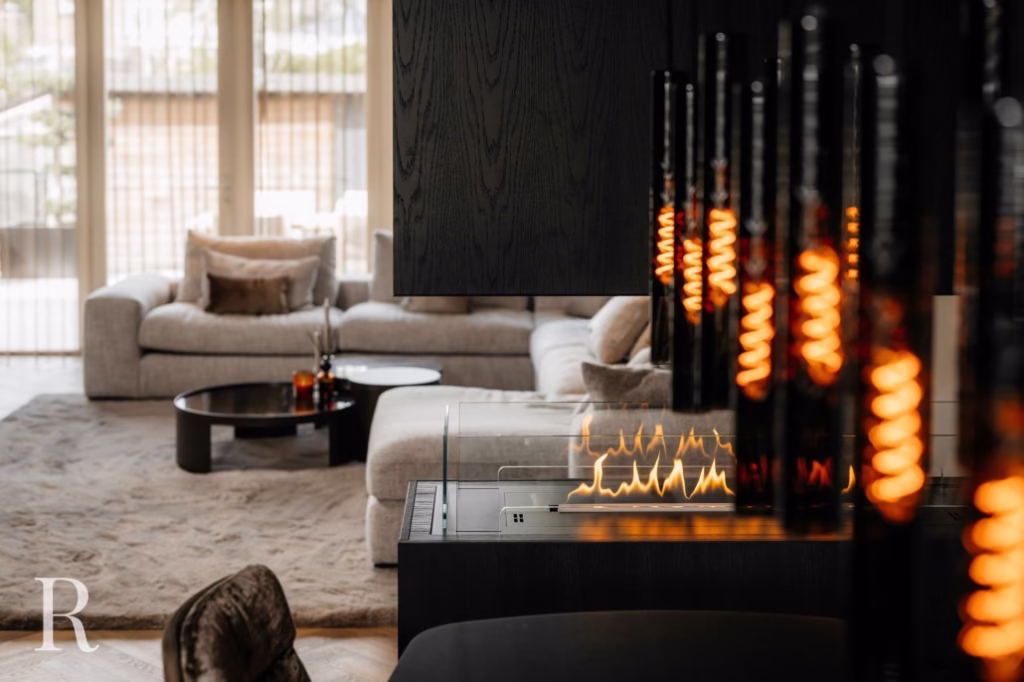 Een moderne woonkamer met een gezellige beige bank, ronde salontafel, pluche vloerkleed en een eigentijdse open haard met zichtbare vlammen en glazen accenten; grote ramen laten natuurlijk licht binnen.