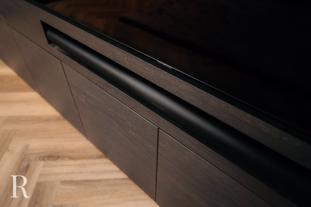 Een close-up van een zwarte Sonos soundbar op een donkerhouten tv-meubel, met een lichte visgraat houten vloer eronder.