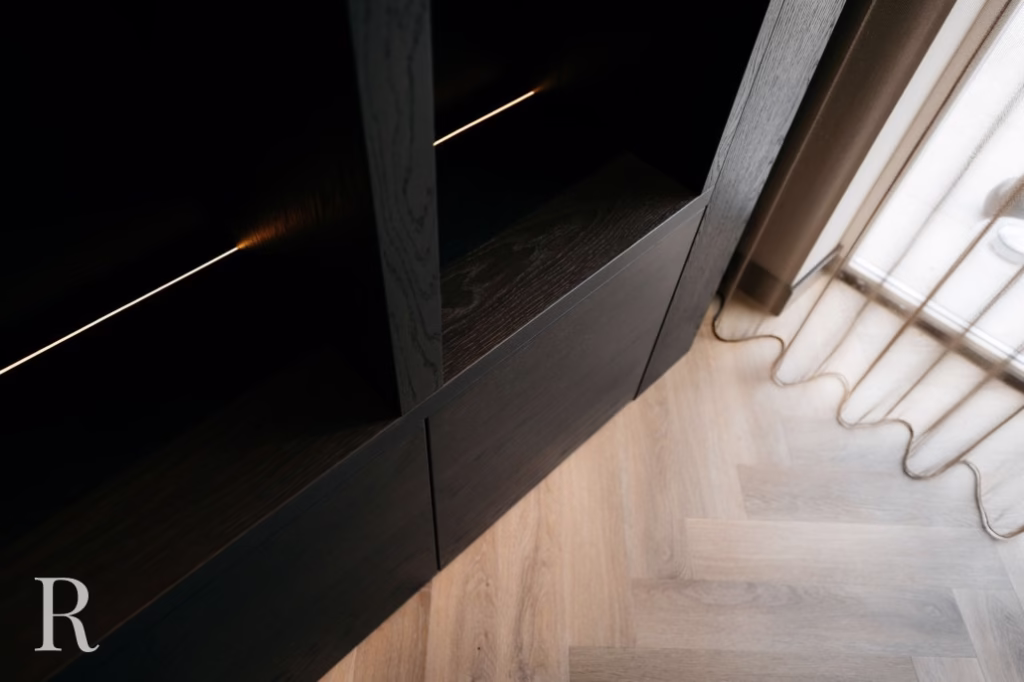 Close-up van een donkere houten kast met open planken en subtiele verlichting. De kast staat op een lichthouten vloer naast een raam met beige gordijnen. Linksonder is een letter R zichtbaar.