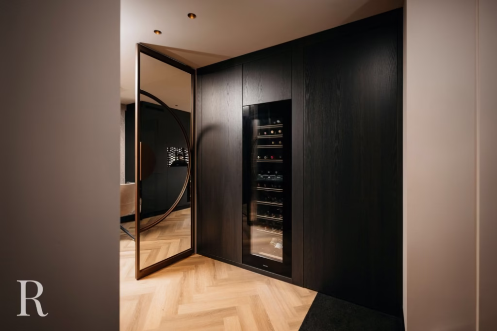 Een moderne wijnkelder met een glazen wijnkoelkast ingebouwd in een donkerhouten muur, een grote ronde spiegeldeur en lichte visgraat houten vloeren. De ruimte is zacht verlicht, wat een luxe gevoel geeft.