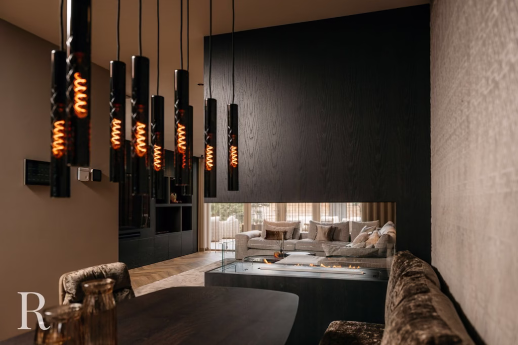 Moderne leefruimte met donkerhouten lambrisering, een ingebouwde glazen open haard en een gezellige zithoek op de achtergrond. Zwarte hanglampen met zichtbare gloeidraden hangen boven een eettafel op de voorgrond.