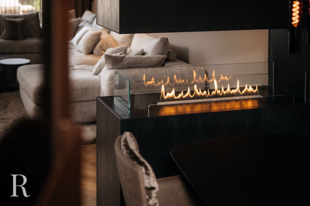 Moderne woonkamer met een lineaire open haard in glas op een donkere console, een beige sofa met kussens op de achtergrond en zachte, warme verlichting die een gezellige sfeer creëert.