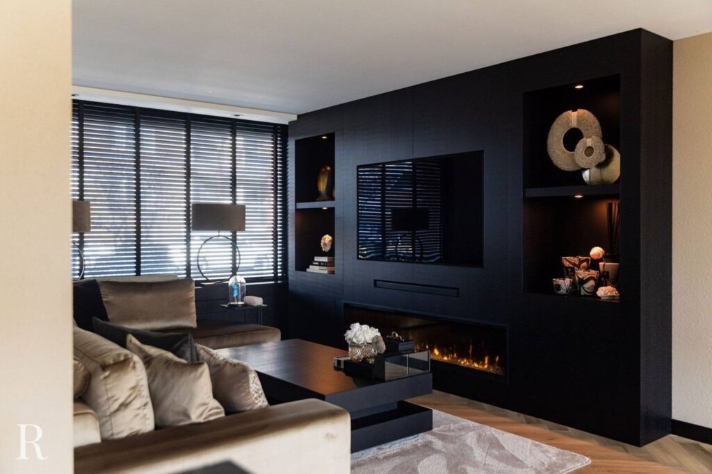Moderne woonkamer met donker ingebouwd wandmeubel met een tv, planken met decor en een lineaire open haard. Neutraal getinte banken, een groot raam met luxaflex, lampen en een vloerkleed maken de elegante, gezellige ruimte compleet.