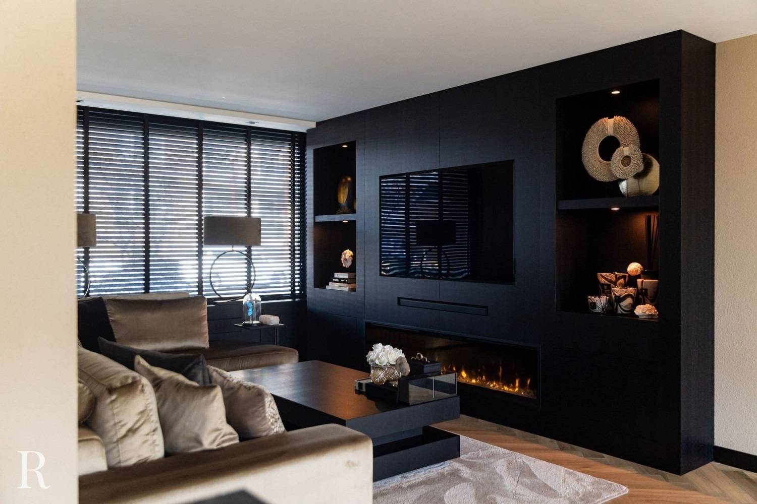 Moderne woonkamer met donker ingebouwd wandmeubel met een tv, planken met decor en een lineaire open haard. Neutraal getinte banken, een groot raam met luxaflex, lampen en een vloerkleed maken de elegante, gezellige ruimte compleet.