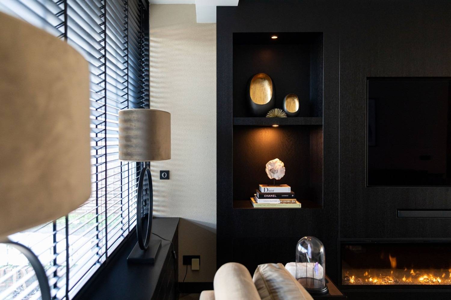 Een moderne woonkamer heeft een zwarte inbouwwand met decoratieve planken met gouden accenten, boeken en een wit beeldhouwwerk. Aan de linkerkant staan een lamp en jaloezieën en onder een aan de muur gemonteerde tv bevindt zich een lineaire open haard.