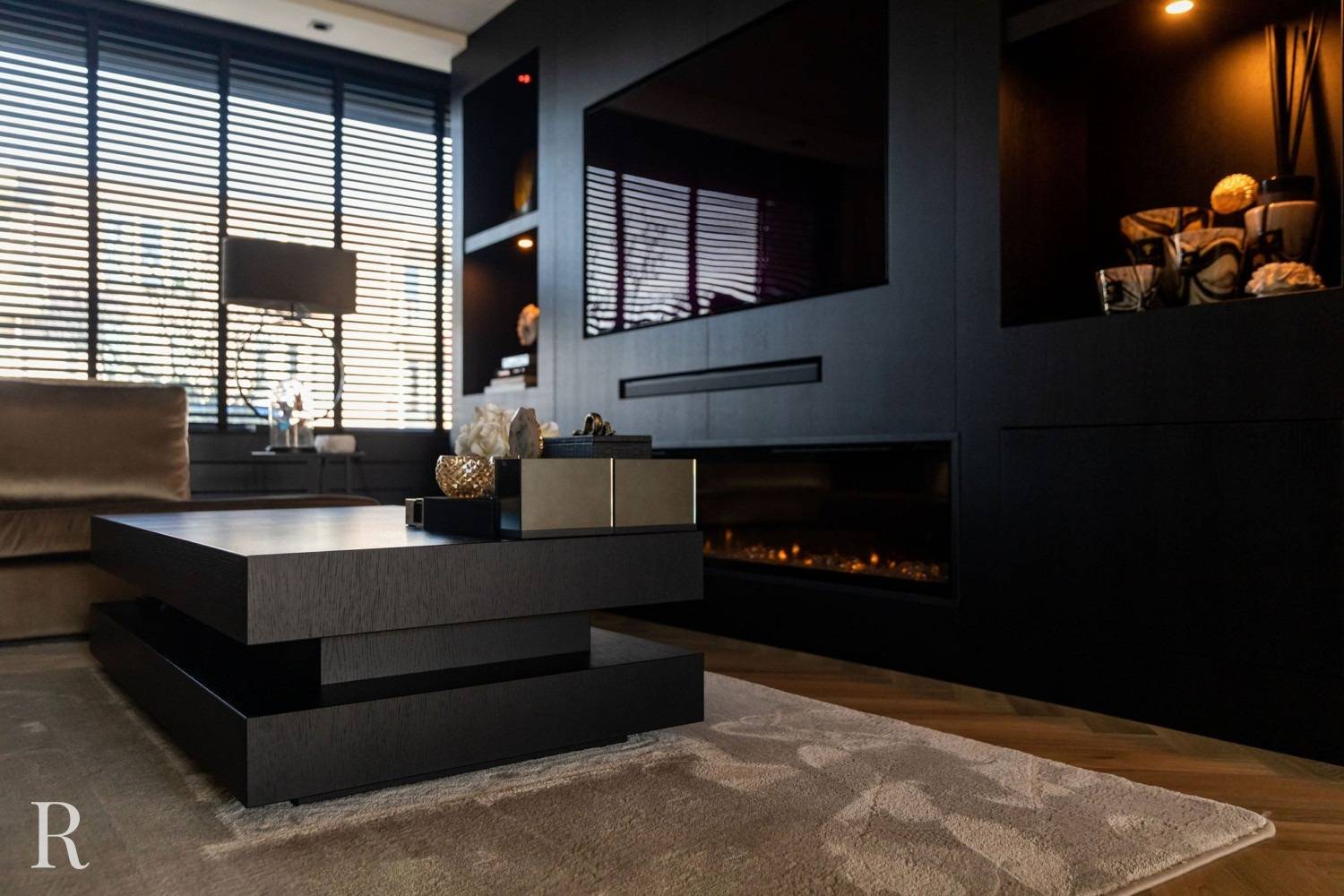 Een moderne woonkamer met een zwarte geometrische salontafel, een beige vloerkleed, een bank, planken met decoratieve voorwerpen, een grote tv en horizontale raamgordijnen die natuurlijk licht binnenlaten. De algehele inrichting is minimaal en eigentijds.