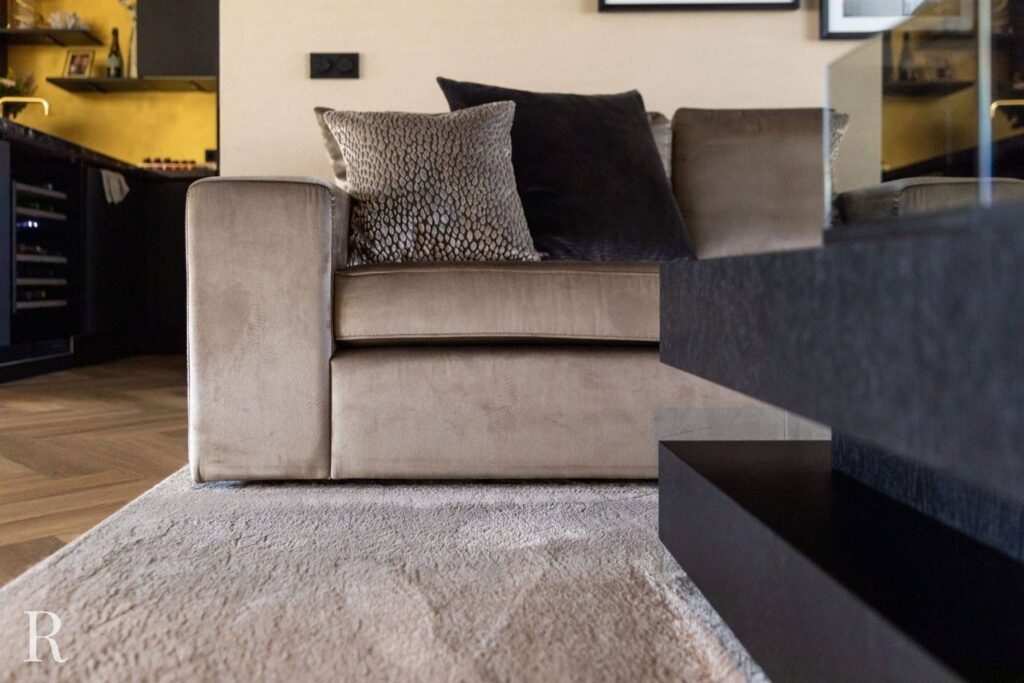 Een moderne woonkamer is voorzien van een beige fluwelen bank met gestructureerde en donkere kussens, geplaatst op een licht vloerkleed. Een deel van een zwarte salontafel en een keuken met warme verlichting zijn zichtbaar op de achtergrond.