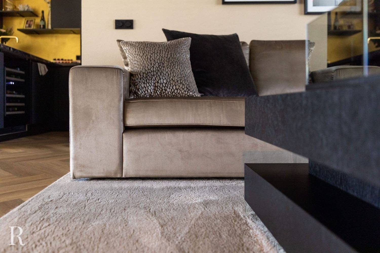 Een moderne woonkamer is voorzien van een beige fluwelen bank met gestructureerde en donkere kussens, geplaatst op een licht vloerkleed. Een deel van een zwarte salontafel en een keuken met warme verlichting zijn zichtbaar op de achtergrond.