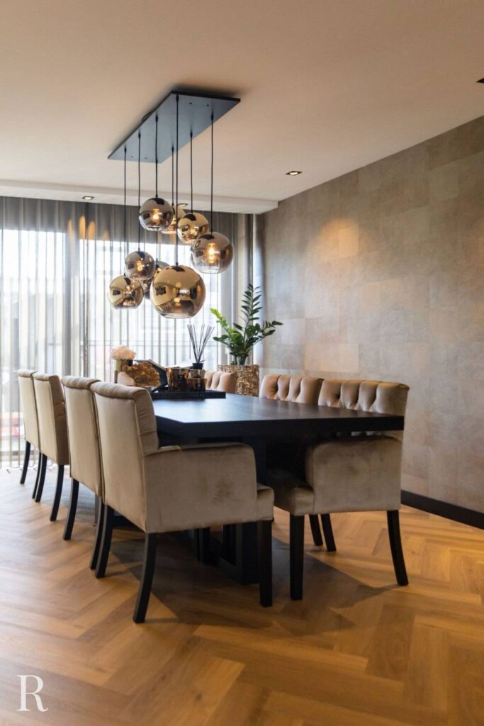 Een moderne eetkamer met een zwarte rechthoekige tafel omringd door acht beige gestoffeerde stoelen, hangende metalen hanglampen erboven, een plantenstuk in het midden en grote ramen met transparante gordijnen.