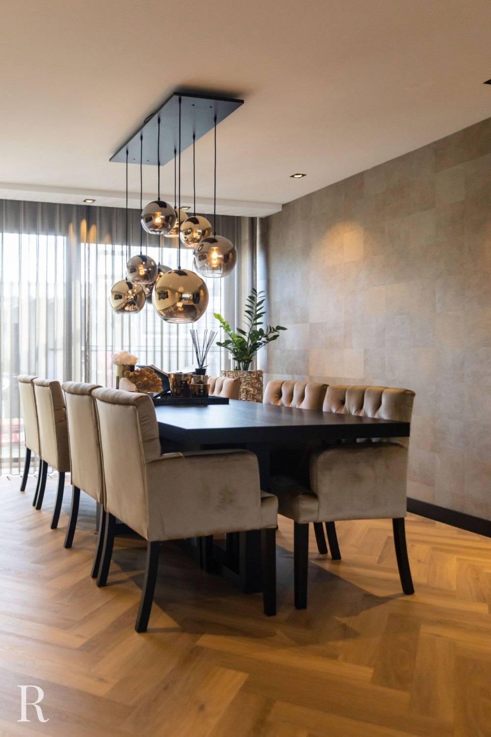 Een moderne eetkamer met een zwarte rechthoekige tafel omringd door acht beige gestoffeerde stoelen, hangende metalen hanglampen erboven, een plantenstuk in het midden en grote ramen met transparante gordijnen.