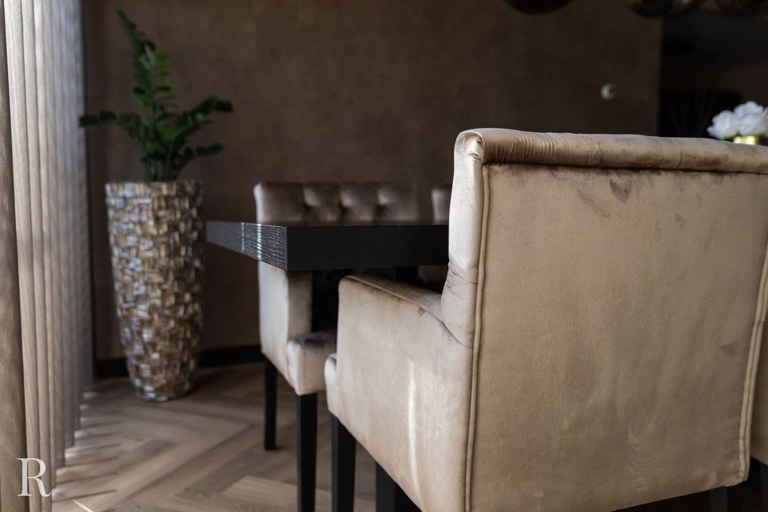 Pluche beige fluwelen eetkamerstoelen omringen een donkere rechthoekige tafel in een moderne kamer met een vloer met houtpatroon. Een hoge decoratieve plant in een vaas met structuur staat in de hoek bij vloerlange gordijnen.