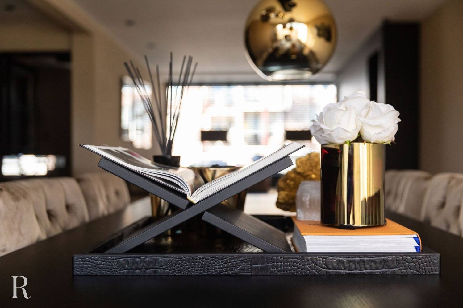 Een modern tafelstuk met een open boek op een zwarte standaard, een gouden vaas met witte rozen en een stapel boeken, allemaal gerangschikt op een donker dienblad met een gouden lamp erboven.