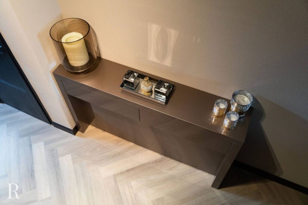 Een moderne consoletafel tegen een beige muur bevat een grote kaars in een glazen houder, een decoratief parfumschaaltje en drie kleine kaarsenhouders. De vloer heeft een licht visgraatmotief.