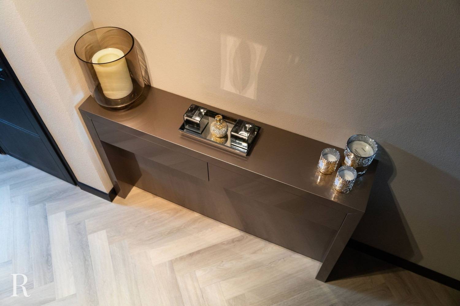 Een moderne consoletafel tegen een beige muur bevat een grote kaars in een glazen houder, een decoratief parfumschaaltje en drie kleine kaarsenhouders. De vloer heeft een licht visgraatmotief.