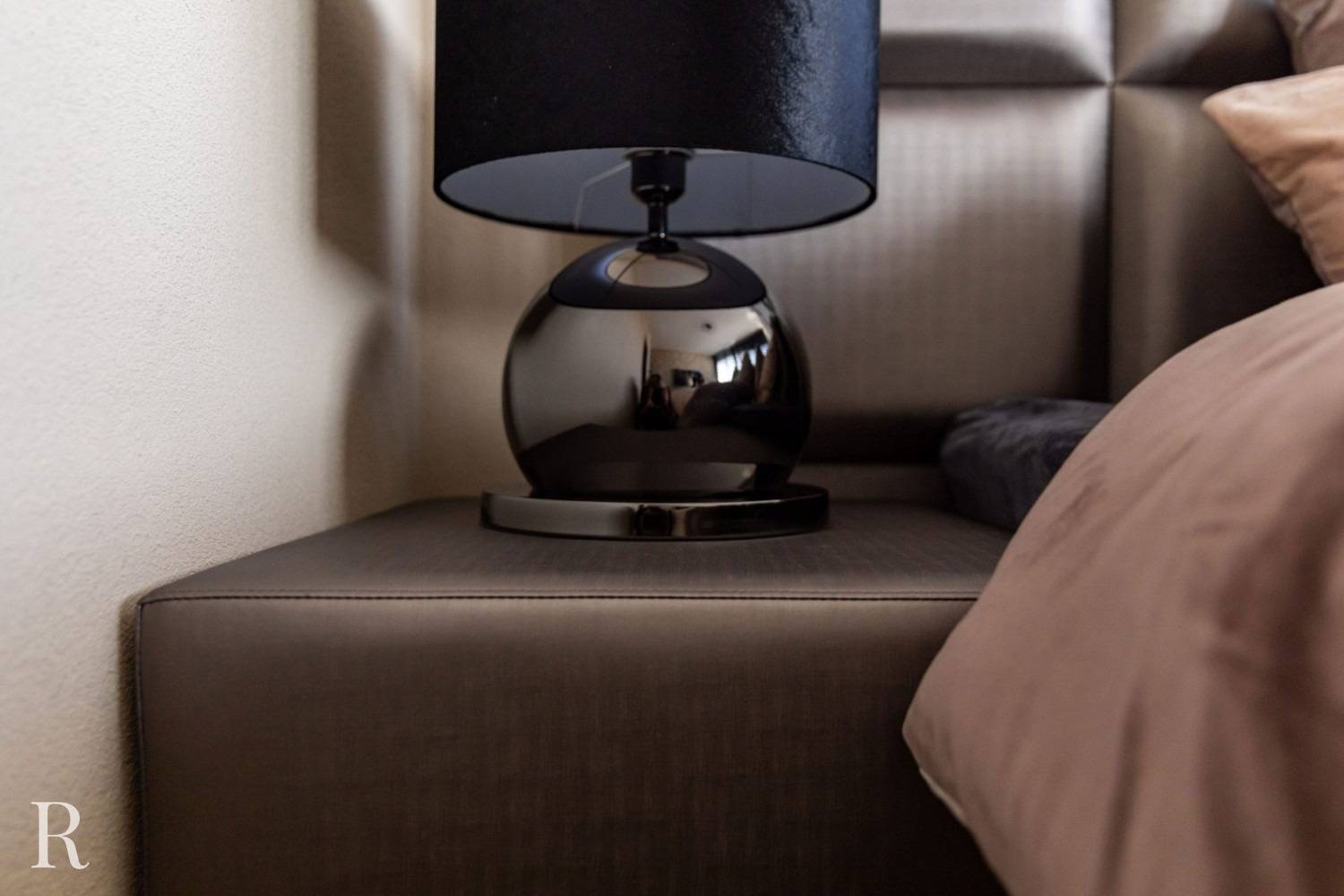 Een zwarte, bolvormige tafellamp met een zwarte lampenkap staat op een bruin nachtkastje naast een bed met bruin en donkergrijs beddengoed. De hoek van de muur en het hoofdeinde zijn zichtbaar.