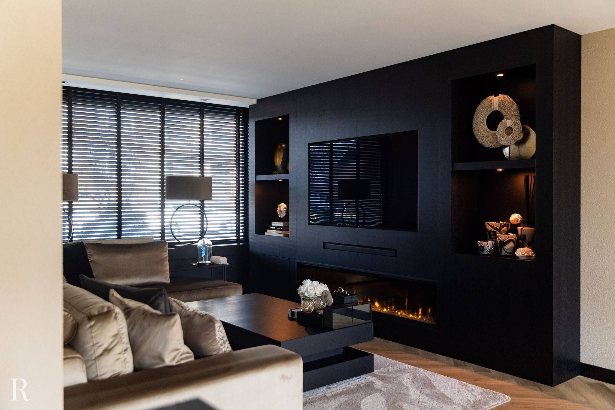 Moderne woonkamer met een zwart ingebouwd wandmeubel met tv, decoratieve planken en elektrische open haard. Neutrale banken, een salontafel, lampen en grote ramen met jaloezieën maken de elegante, gezellige ruimte compleet.