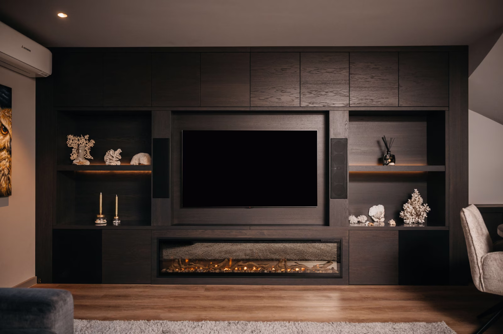 Een moderne woonkamer heeft een donkerhouten entertainmentcentrum met planken met decoratieve voorwerpen, een flatscreentelevisie in het midden en een elektrische open haard eronder, allemaal op een houten vloer.