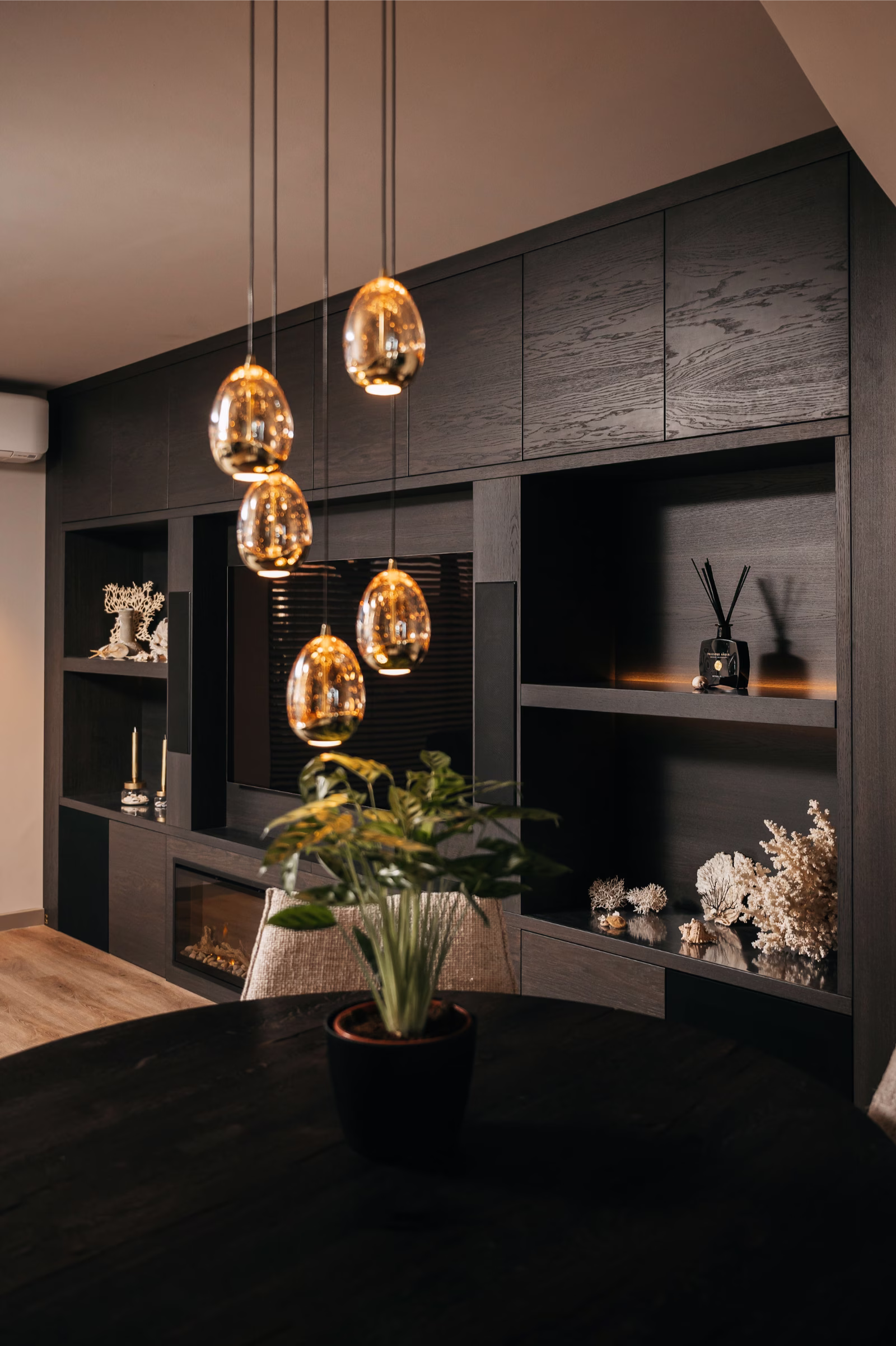 Moderne leefruimte met donkerhouten planken, decoratieve voorwerpen en een potplant op een ronde tafel. Verschillende warme, hangende hanglampen zijn boven de tafel geplaatst en creëren een gezellige sfeer.